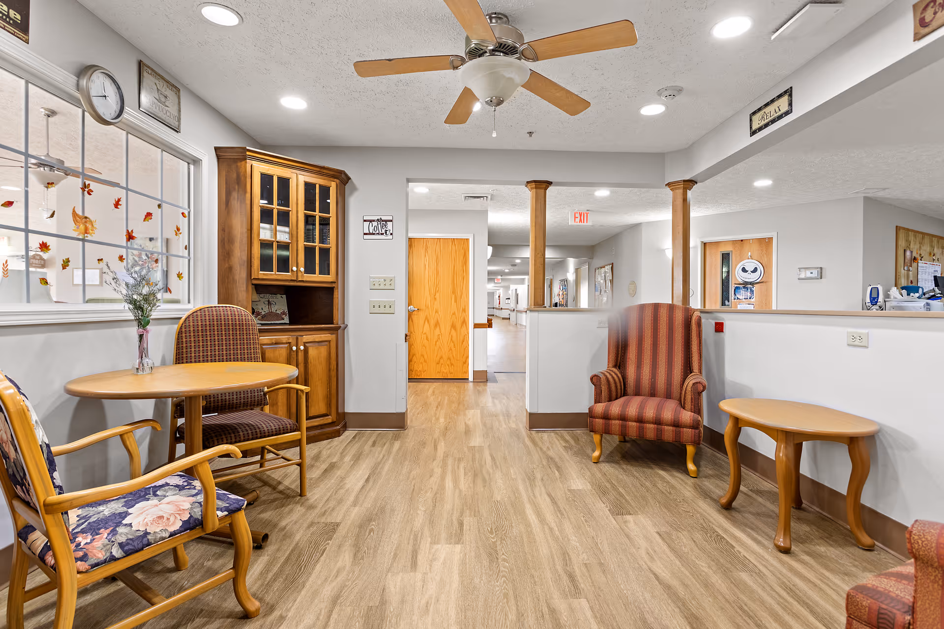 A cozy seating area in a senior living facility with wooden flooring, a ceiling fan, and recessed lighting. The area features a round wooden table with two chairs, a wooden cabinet with glass doors, and a striped armchair next to a small wooden side table. The walls are light gray, and there is a window decorated with autumn leaf decals. A hallway with wooden doors and an exit sign is visible in the background.