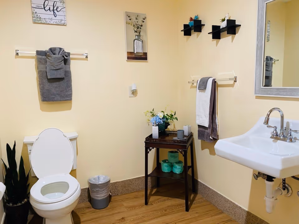 A clean and well-lit bathroom with a white toilet with the lid open, a small trash can beside it, and a white sink with a silver faucet. There are gray and white towels hanging on towel racks on the walls. A small wooden table holds decorative items including a vase with flowers and some rolls of toilet paper. The walls are painted light yellow and decorated with framed pictures and small black shelves with plants.