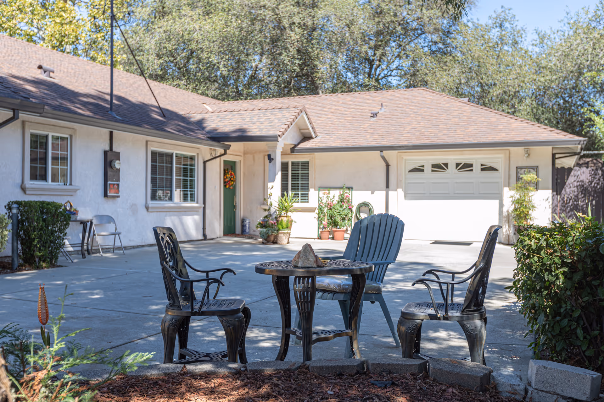 Outdoor patio area at Citrus Pines Senior Living with a round metal table and three chairs on a concrete surface. The background shows a single-story building with a green door, windows, potted plants, and a garage door. Trees and bushes surround the area under a clear sky.