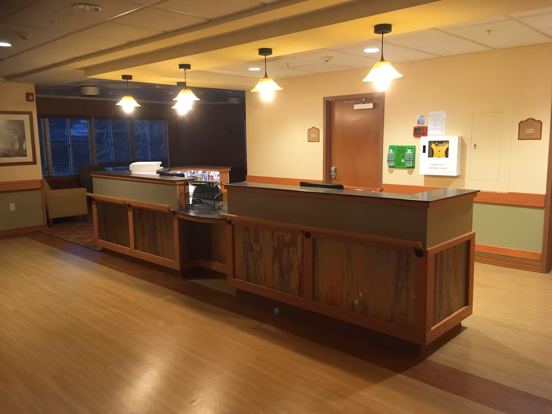 A reception desk in a lobby with pendant lights, wood floors, and seating near windows.
