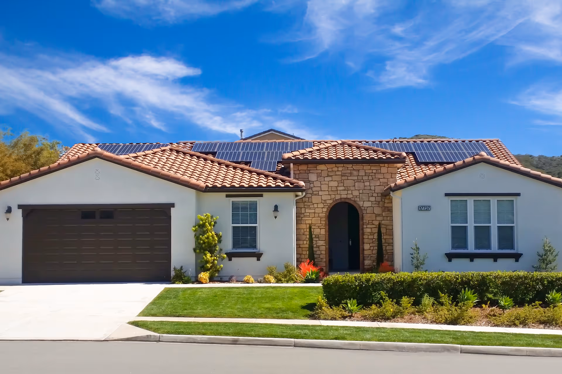 Front exterior of a single-story stucco house with a tile roof, garage, solar panels, and a landscaped lawn.