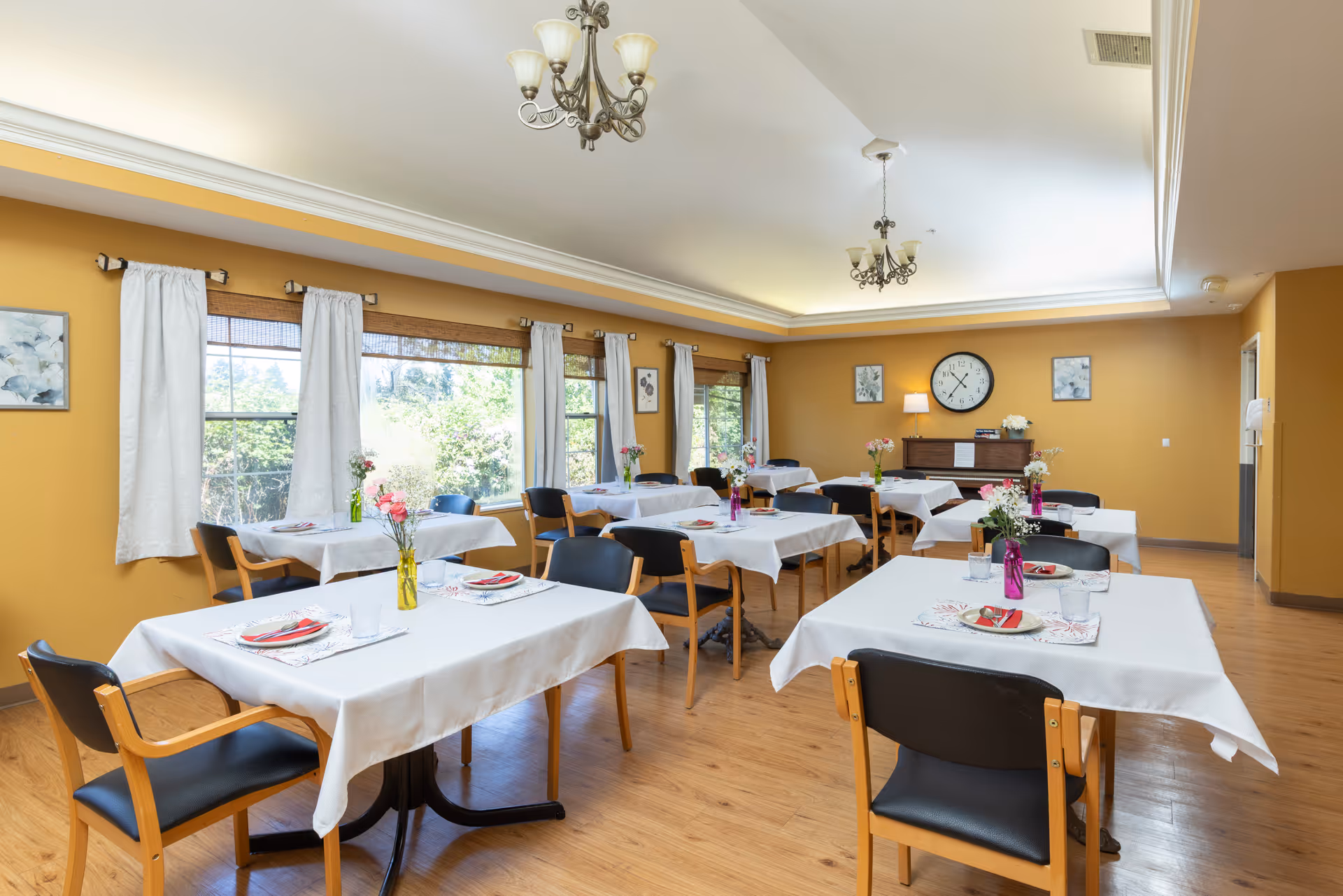 Bright dining room with multiple tables set with white tablecloths, chairs, floral centerpieces, large windows, and a clock on the far wall.