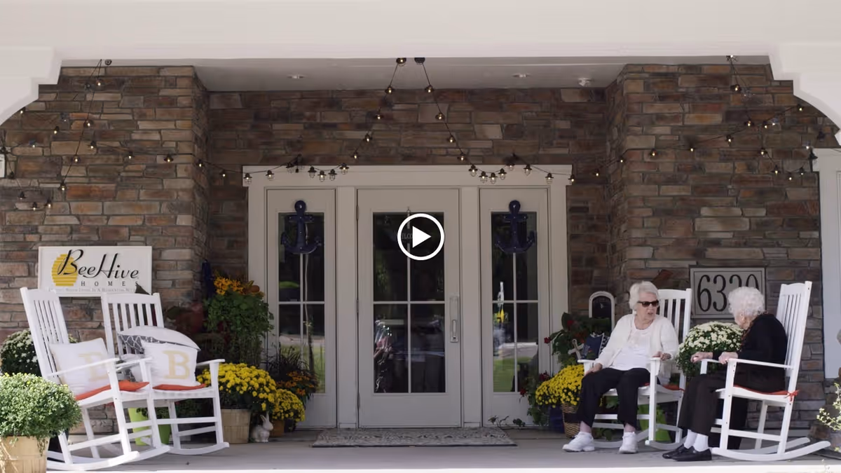 Entrance porch of BeeHive Homes Assisted Living with rocking chairs, potted flowers, and two women seated near the front doors.