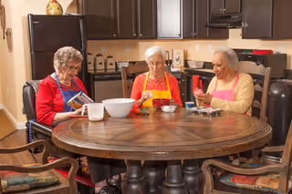 Three elderly women sitting around a round wooden table in a kitchen preparing food together.