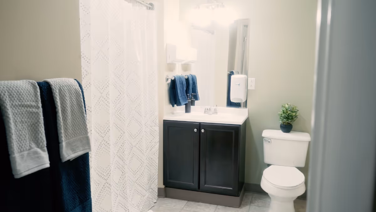 Bathroom with a dark wood vanity and sink, mirror, toilet with a small potted plant, shower with a white patterned curtain, and blue towels.
