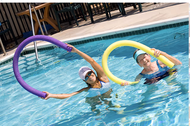Two women in a swimming pool holding colorful pool noodles, smiling and engaging in water exercise near the pool edge with chairs and a fence in the background.