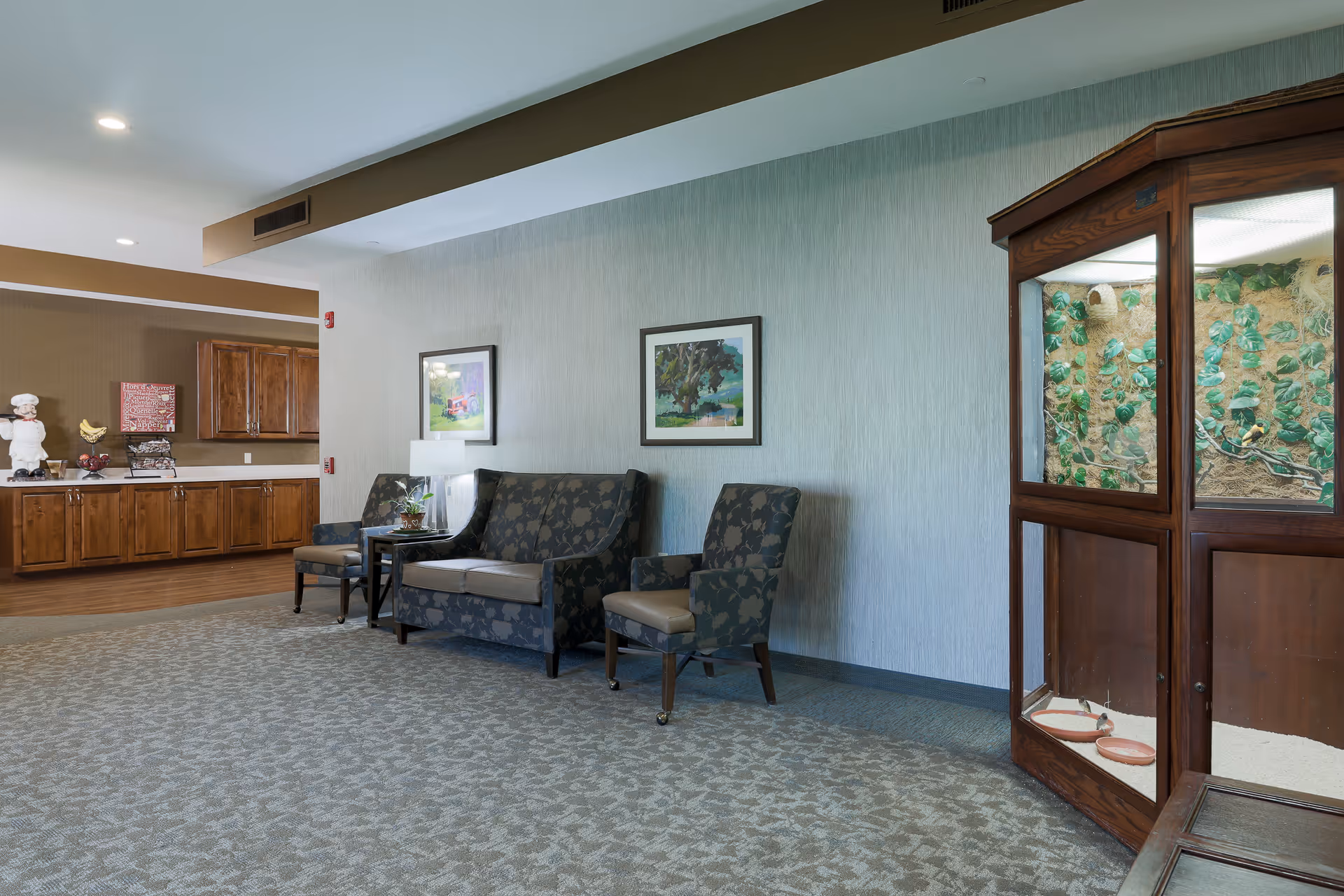 A seating area in a senior living facility with two patterned armchairs and a matching loveseat arranged around a small table with a lamp and a plant. The wall behind the seating area has two framed landscape paintings. To the right, there is a large wooden enclosure with glass panels containing a decorative setup with plants and birds. In the background, there is a wooden cabinet with various decorative items on top, including a chef figurine and a banana sculpture.