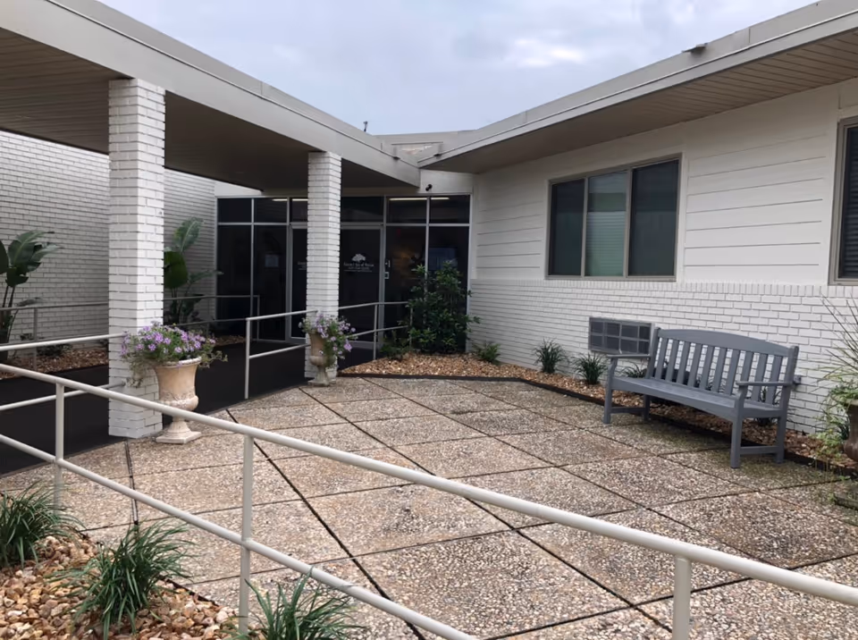 Entrance courtyard of a senior living facility with a covered walkway, accessibility ramp, potted flowers, and a bench by a white brick building.