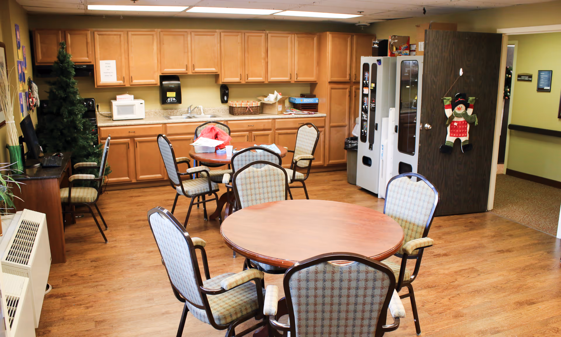 Communal dining area with round tables and chairs, a kitchenette with cabinets and appliances, and a vending machine.