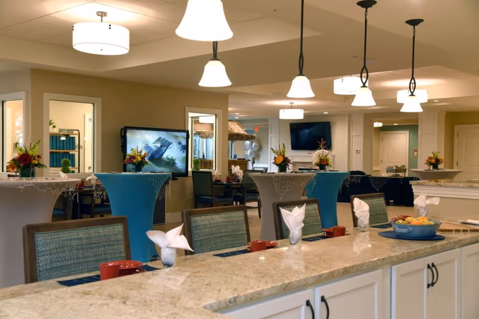 Interior view of a senior living facility dining area with a long granite countertop in the foreground set with napkins and cups. Behind the counter are several high tables covered with blue and white tablecloths and decorated with flower arrangements. The room is well-lit with hanging pendant lights and ceiling fixtures. In the background, there are chairs, a TV mounted on the wall, and doorways leading to other rooms.