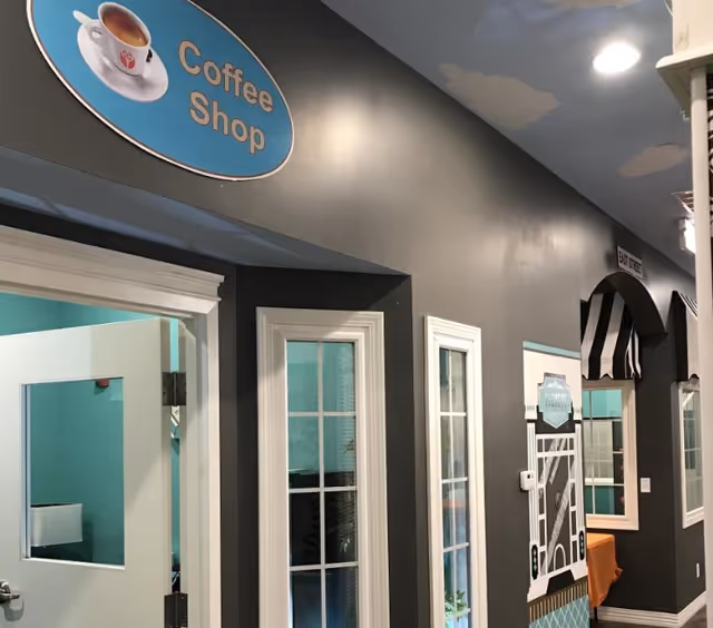 Interior hallway of a facility with a dark gray wall featuring a sign that says 'Coffee Shop' above a door. The hallway has windows with white frames and decorative elements resembling storefronts, including a black and white striped awning and a sign that says 'Exit Here'. The ceiling is painted with clouds on a blue background.