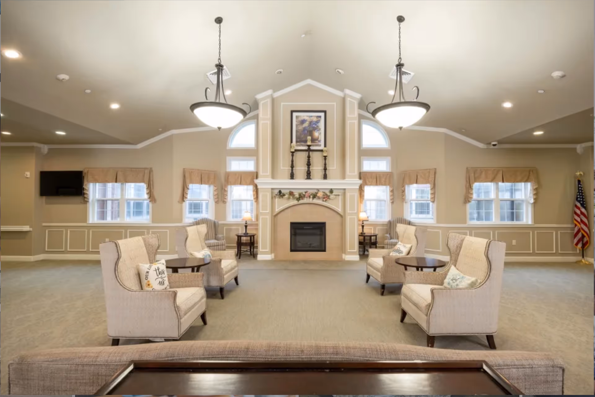 A spacious and well-lit living room area with beige walls and carpet. The room features a central fireplace with decorative candles and a framed picture above it. There are two large hanging light fixtures on the ceiling. Four upholstered armchairs with patterned cushions are arranged in pairs facing each other with small round tables between them. Several windows with beige valances allow natural light to enter. An American flag stands in the corner on the right side of the room.