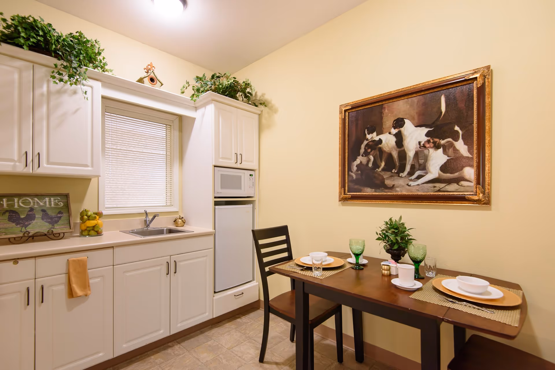 A small kitchen and dining area with white cabinets, a sink under a window with blinds, a microwave above a mini refrigerator, and a wooden dining table set for two with green glasses and white dishes. There is a framed painting of dogs on the wall and green plants on top of the cabinets.