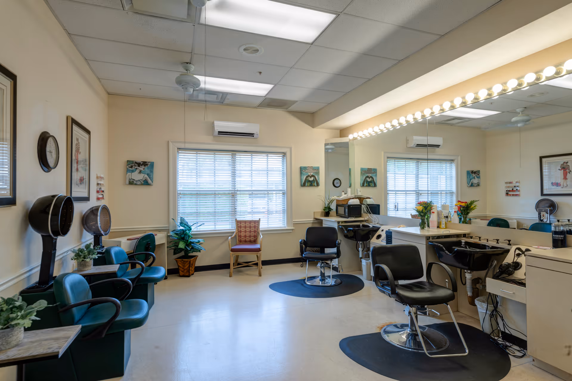 Interior view of a hair salon area in a senior living facility with salon chairs, hair dryers, a large mirror with lights, plants, and framed artwork on the walls. There is a window with blinds and a ceiling fan above.