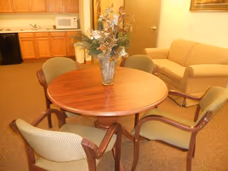 A cozy interior room featuring a round wooden table with a floral centerpiece surrounded by four cushioned chairs. In the background, there is a beige sofa and a kitchenette area with wooden cabinets, a microwave, and a small refrigerator.