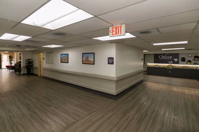Interior view of a senior living facility hallway with wood-look flooring, white walls, and ceiling lights. There are framed pictures on the walls, a reception desk with the name 'Osborn' visible, and an exit sign hanging from the ceiling.