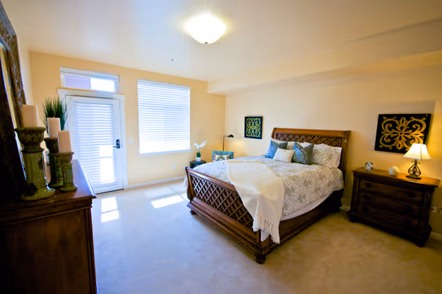 A bright bedroom with a large wooden bed featuring a patterned bedspread and multiple pillows. There is a wooden nightstand with a lamp and decorative items on the right side of the bed. On the left side, there is a dresser with candles and a plant. The room has beige walls, a window with blinds, and a glass door letting in natural light.