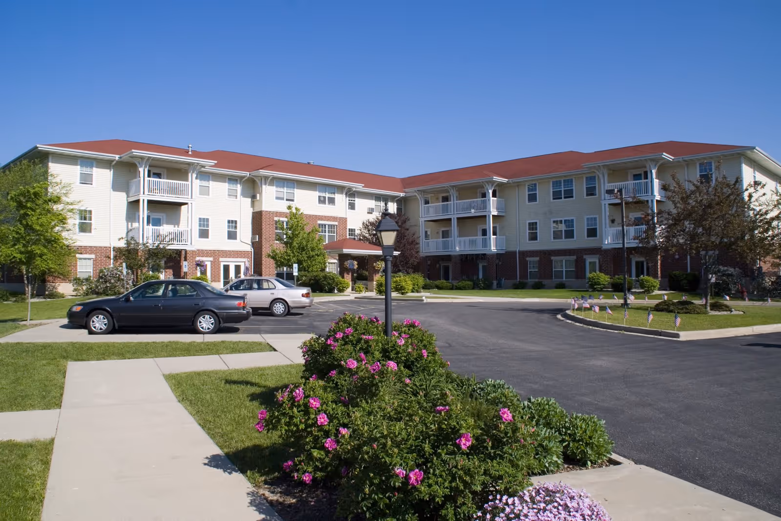 Exterior view of Bell Tower Place Senior Apartments showing a three-story building with beige siding and red roof. There are balconies on the second and third floors, a parking lot with cars, a sidewalk, and landscaped bushes with pink flowers in the foreground under a clear blue sky.
