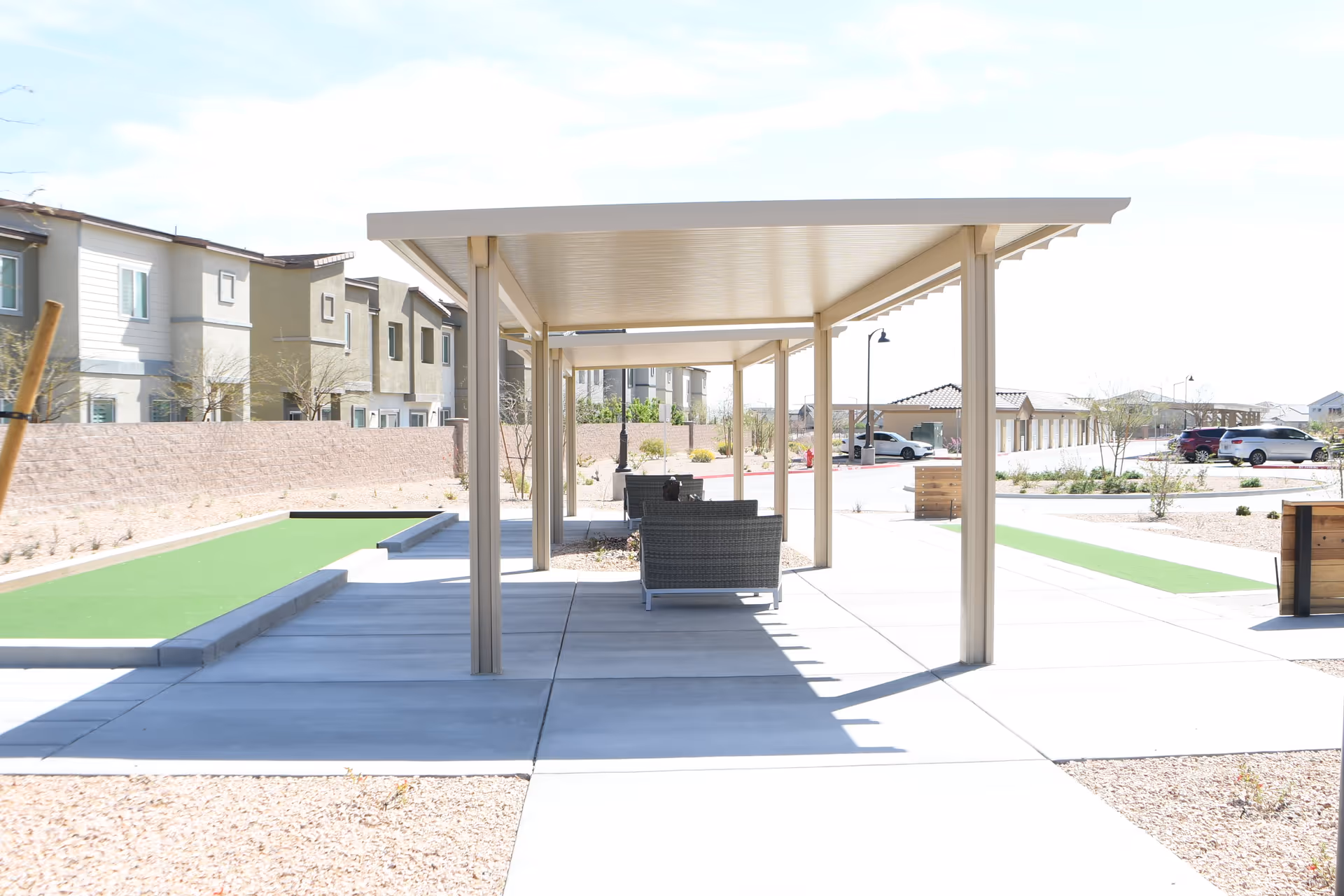 Outdoor covered seating area with wicker chairs under a beige pergola, adjacent to a bocce ball court, with residential buildings and parked cars in the background under a clear sky.