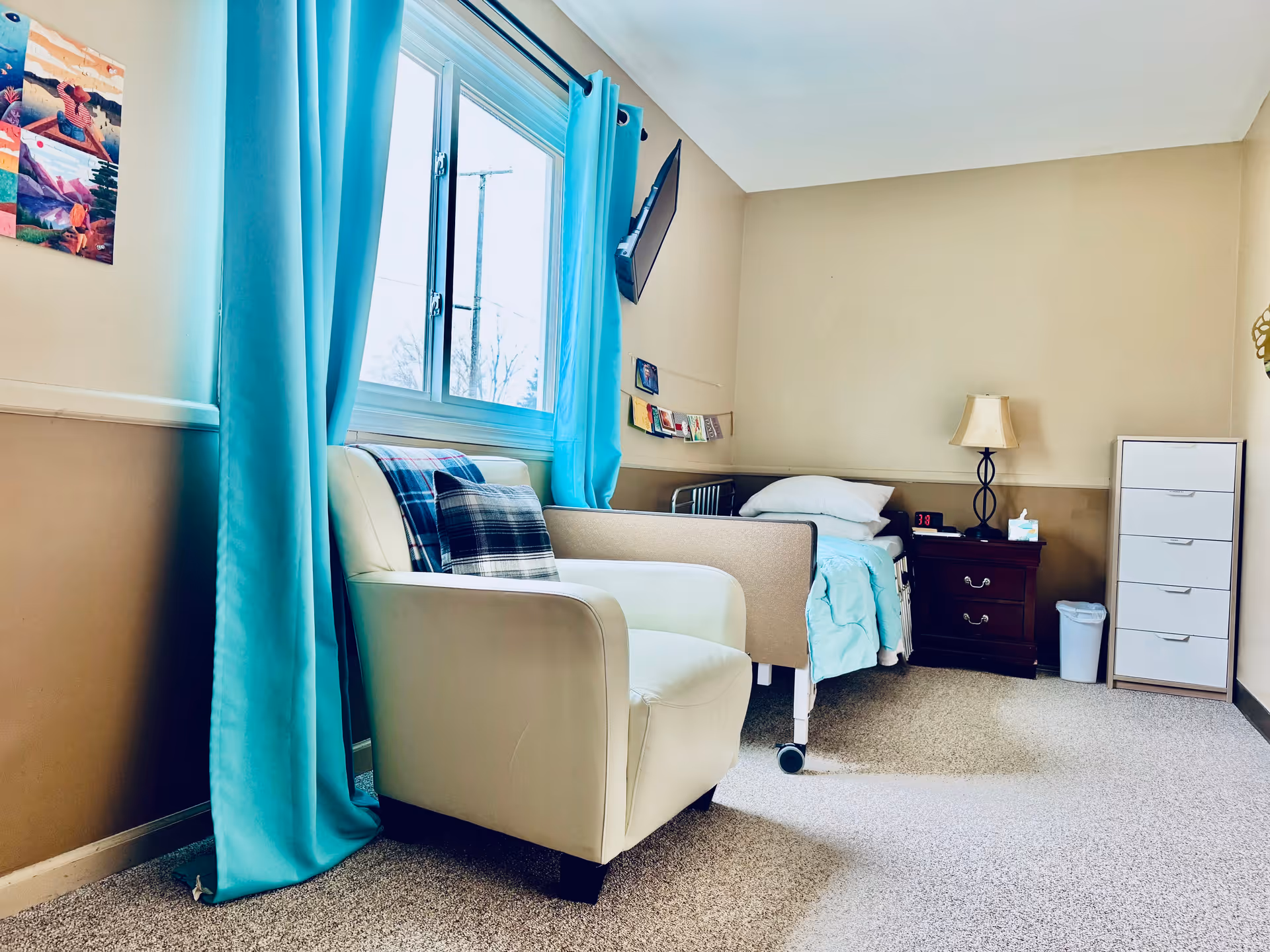 A cozy bedroom in Aiello Adult Foster Care featuring a hospital-style bed with light blue bedding, a white armchair with plaid pillows, a window with bright turquoise curtains, a wall-mounted TV, a dark wooden nightstand with a lamp and tissue box, a white dresser, and beige walls with carpeted floor.