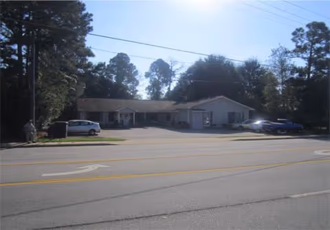 Single-story building with a light-colored exterior situated behind a paved road with yellow dividing lines. Several cars are parked in front of the building, which is surrounded by tall trees under a clear blue sky.