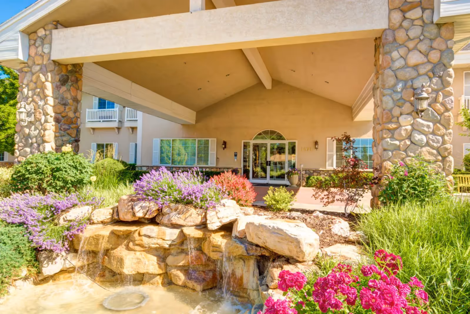 Covered building entrance with stone pillars, a landscaped rock waterfall, and colorful flower beds.