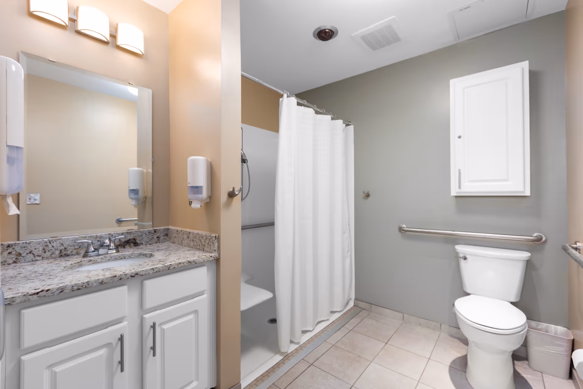 A clean and accessible bathroom with a granite countertop sink, white cabinets, a large mirror, a wall-mounted soap dispenser, a walk-in shower with a white curtain and built-in seat, a toilet with grab bars on the walls, and a small trash can on the tiled floor.