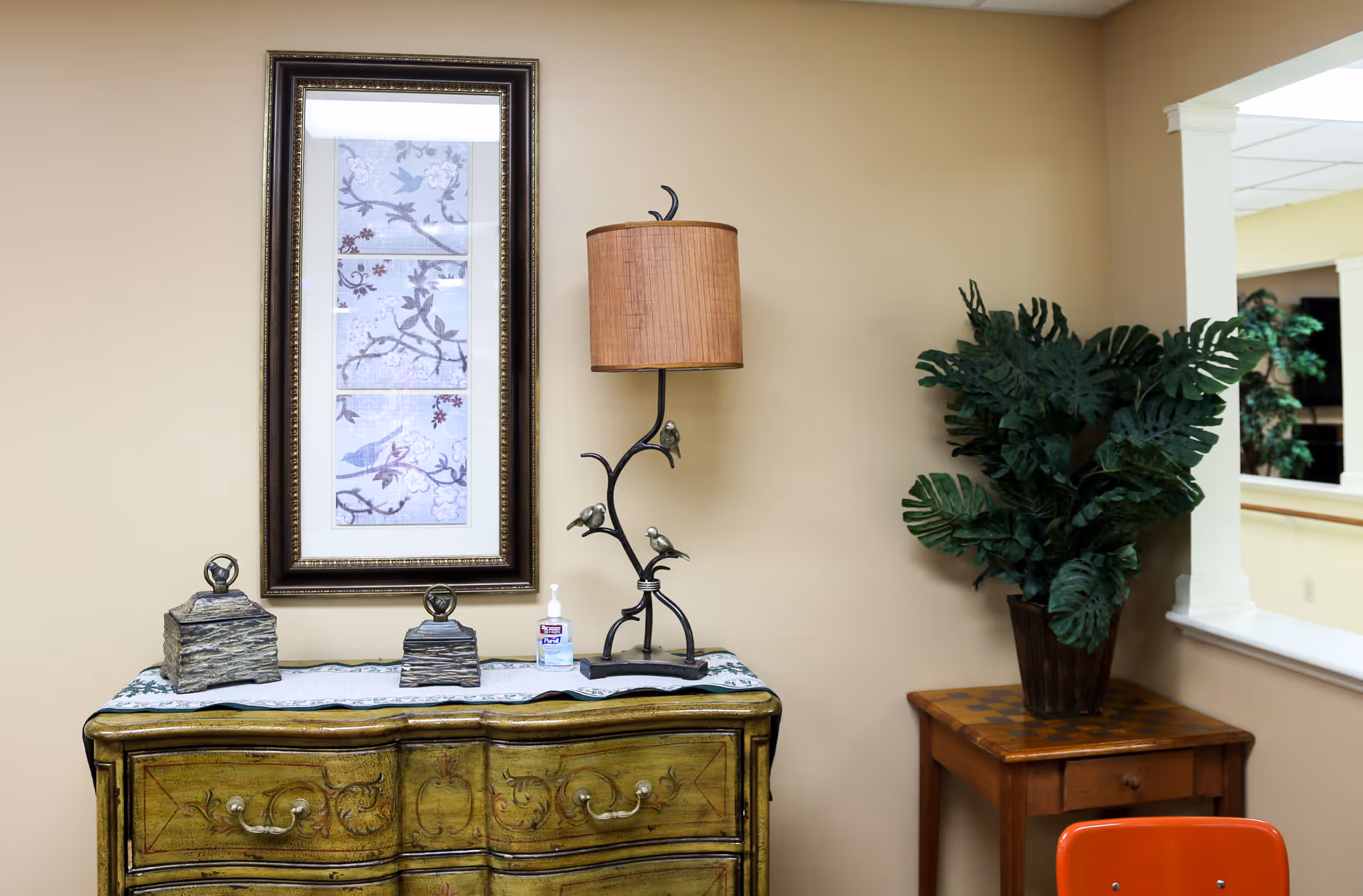 An interior corner of a room with a decorative greenish-yellow chest of drawers topped with two small decorative boxes, a bottle of hand sanitizer, and a lamp with a bird design. Above the chest is a framed artwork featuring birds and branches. To the right, there is a small wooden table with a large potted plant. The walls are beige, and there is a window opening to another room with more plants visible.
