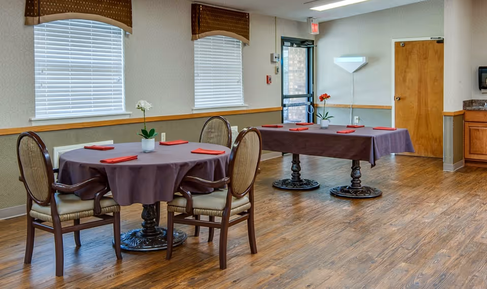 A dining area with two tables covered in purple tablecloths, each set with red napkins and a small flower vase. The room has wooden flooring, two windows with blinds and brown valances, a door with a glass panel, and a wooden door. There is a wall-mounted light fixture and a countertop with cabinets on the right side.