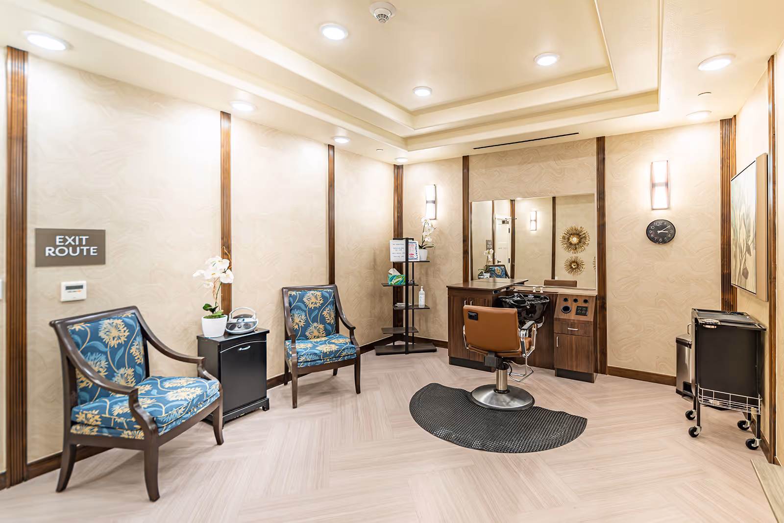 A well-lit salon area in a senior living facility with a single brown salon chair in front of a large mirror and wash basin. Two blue patterned armchairs are placed against the wall near a small black cabinet with a white orchid plant and a radio. A black mat is on the floor beneath the salon chair. The walls are beige with wooden vertical accents, and there is a clock and decorative wall art on the right side. An exit route sign is visible on the left wall.