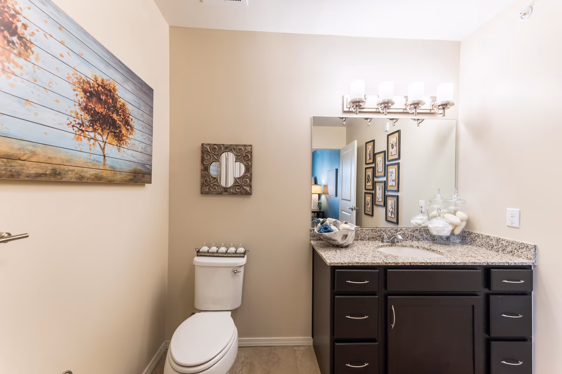 A clean and well-lit bathroom with beige walls, featuring a white toilet with decorative items on the tank lid, a dark wood vanity with a granite countertop, a sink, and a large mirror above it. Above the mirror is a modern light fixture with five lights. On the wall to the left, there is a framed artwork of a tree with autumn leaves, and a decorative mirror is hung above the toilet.