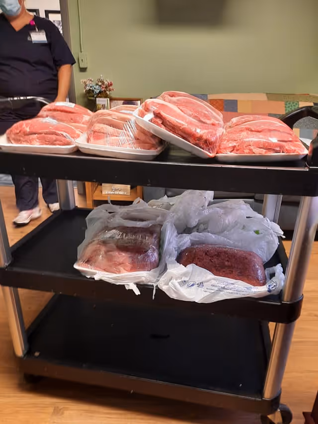 A black utility cart with multiple packages of raw meat on two shelves inside a room with wooden flooring. A person wearing dark scrubs and a face mask is partially visible standing behind the cart. In the background, there is a couch with a colorful patchwork quilt and a small table with a flower arrangement.