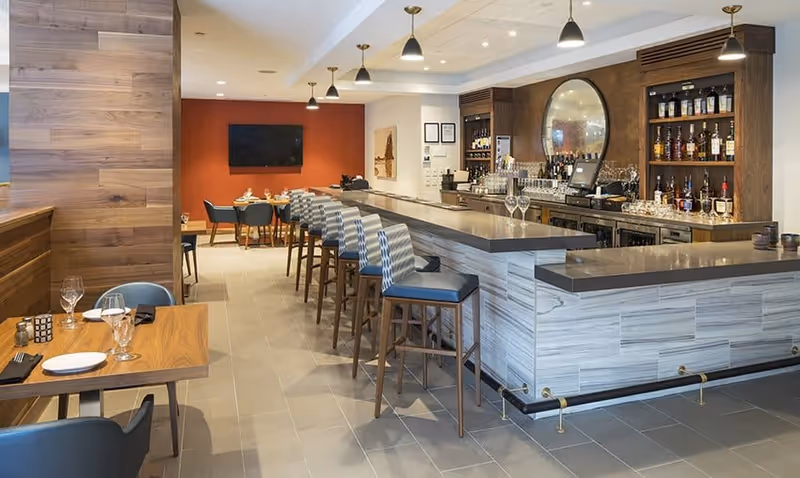 A modern bar and dining area with a long counter lined with high chairs, a wooden wall on the left, tables set with plates and glasses, and shelves stocked with bottles behind the bar.