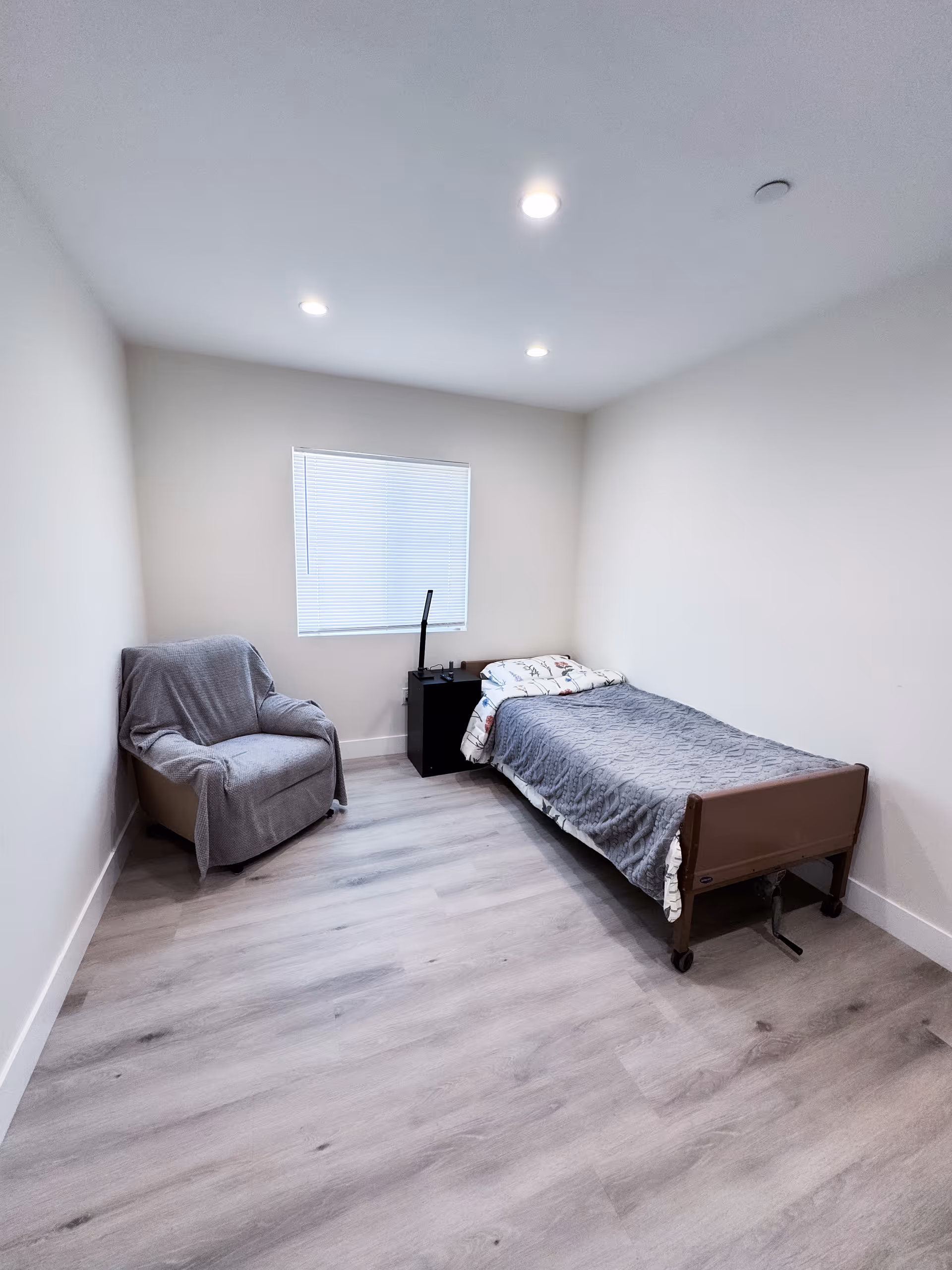 A small, simple bedroom with a single bed covered with a gray quilt and a patterned blanket, a gray armchair draped with a gray cover, a black nightstand with a lamp, and a window with closed blinds. The room has light-colored walls and wood flooring with recessed ceiling lights.