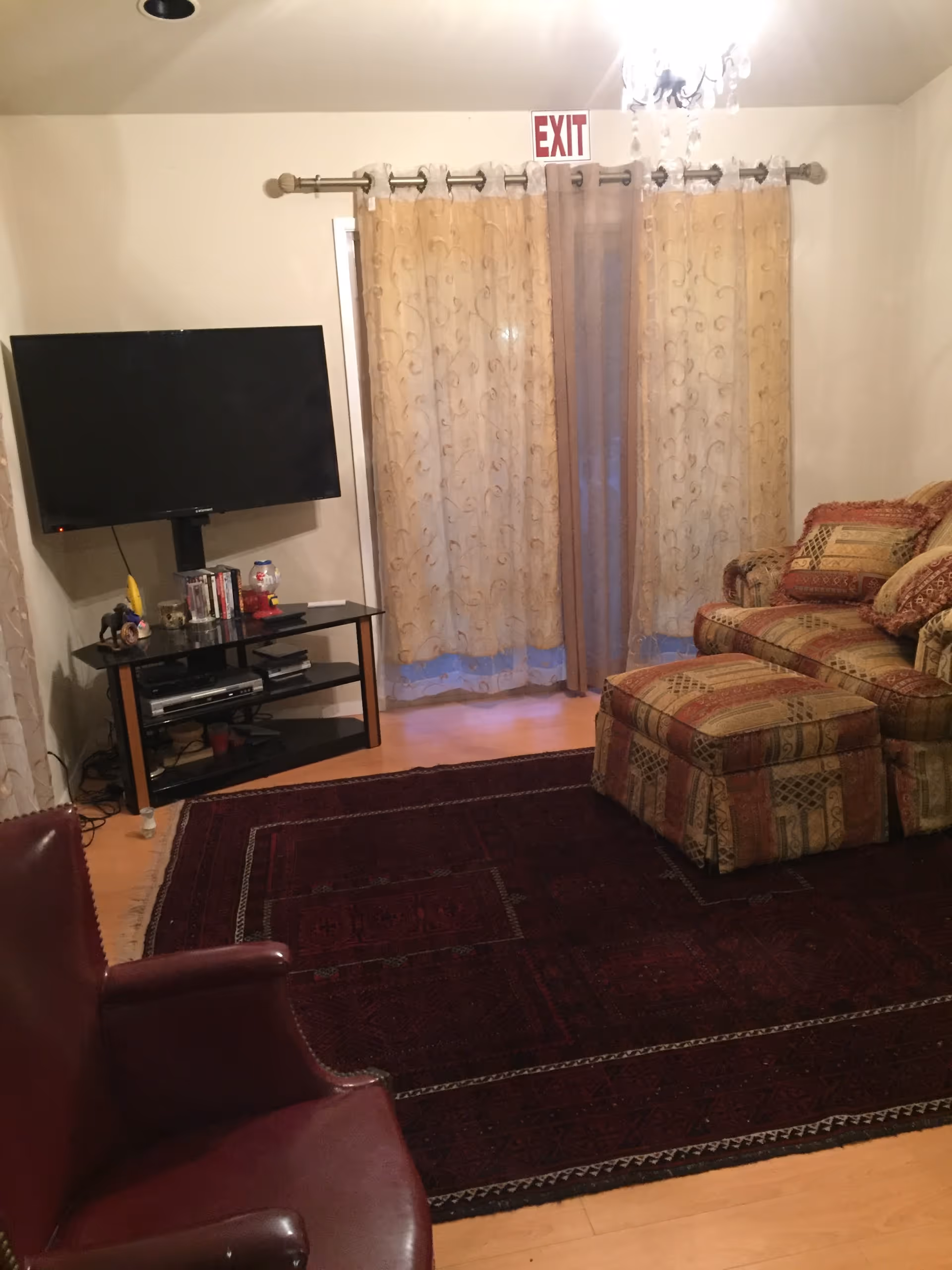 Living room with a patterned armchair and ottoman, wall-mounted TV on a stand, sheer curtains beneath an EXIT sign, and a dark area rug on hardwood floor.