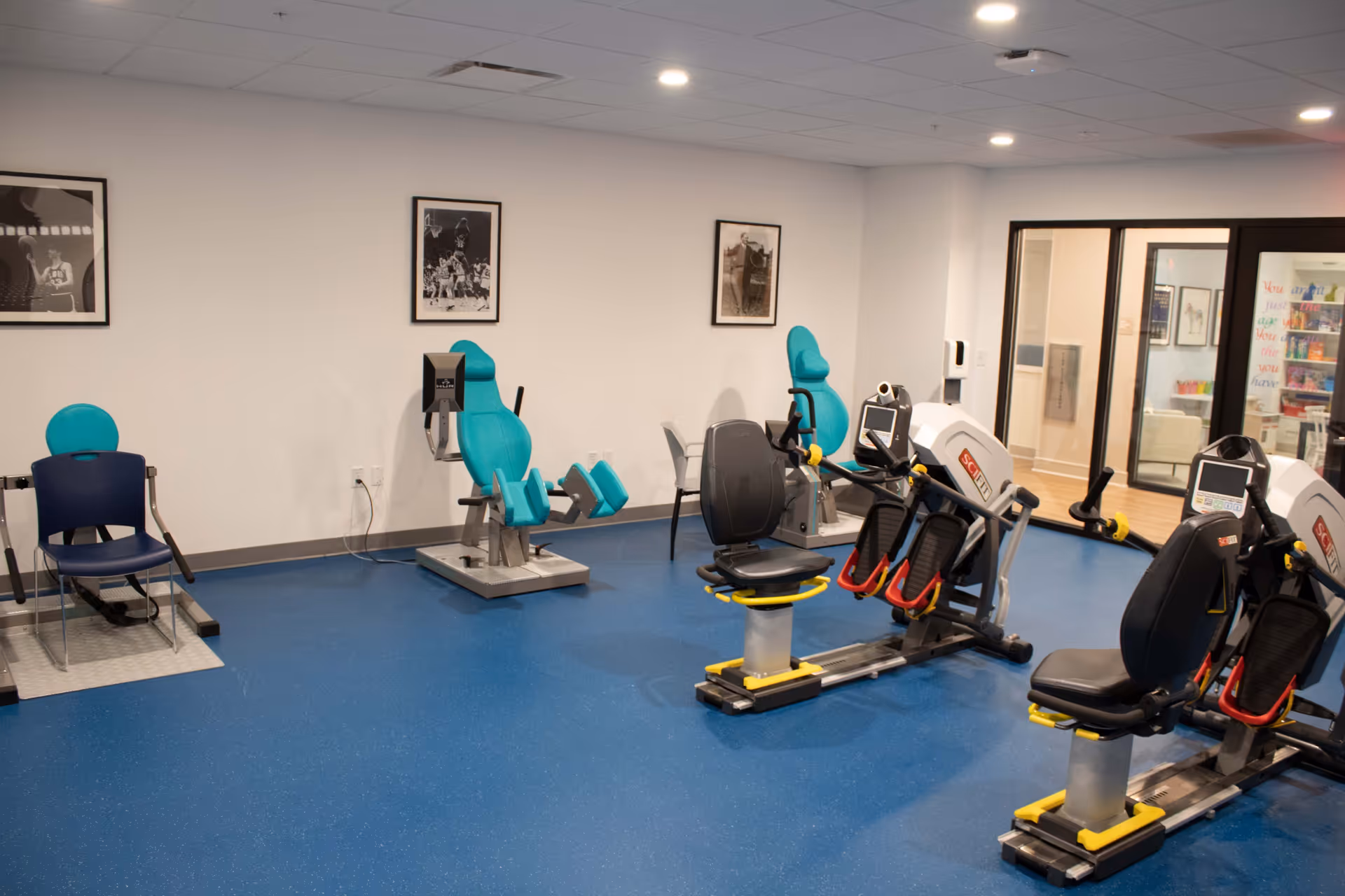 A fitness room with exercise equipment including two recumbent bikes, two leg exercise machines with teal seats, and a blue chair. The room has a blue floor, white walls with three framed black and white photos, and a glass door leading to another room.