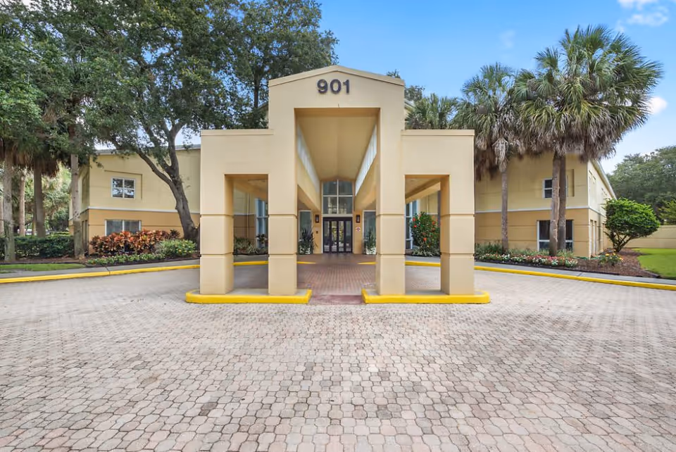 Front exterior view of a beige two-story building with the number 901 above the entrance, surrounded by palm trees and greenery under a blue sky with some clouds.