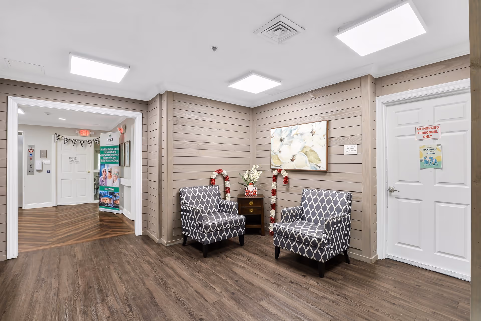A small seating area in a senior living facility with two patterned armchairs facing each other, a small wooden side table with a vase of flowers between them, and a floral painting on the wall. The walls are paneled with light wood, and the floor has dark wood flooring. There is a white door with a sign that reads 'AUTHORIZED PERSONNEL ONLY' and a hallway leading to another area with a banner and informational stand.