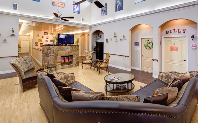 A cozy assisted living facility common area with a large brown leather sectional sofa, patterned armchairs, a round glass coffee table, and a stone fireplace with a mounted flat-screen TV above it. The room has light wood flooring, white walls with decorative arches, and ceiling fans. There are doors labeled with names and numbers, and various decorations including wall art and small shelves with figurines.