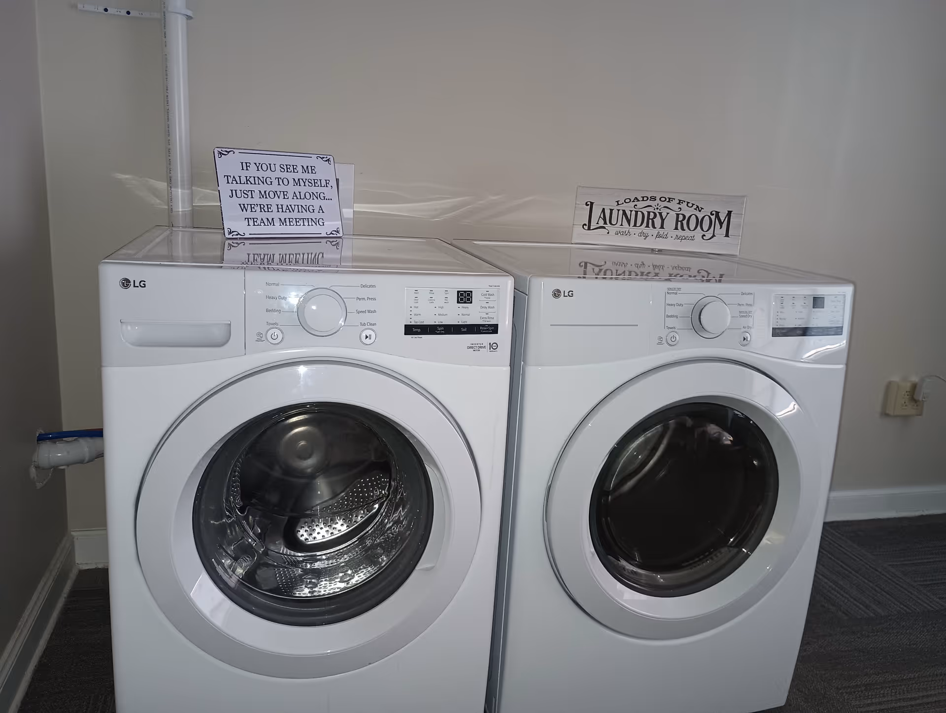 Two white LG front-loading laundry machines, a washer and a dryer, placed side by side against a beige wall. On top of the washer is a small sign that reads 'IF YOU SEE ME TALKING TO MYSELF, JUST MOVE ALONG... WE'RE HAVING A TEAM MEETING.' On top of the dryer is a sign that reads 'LOADS OF FUN LAUNDRY ROOM wash - dry - fold - repeat.' The floor is carpeted with a dark patterned carpet.