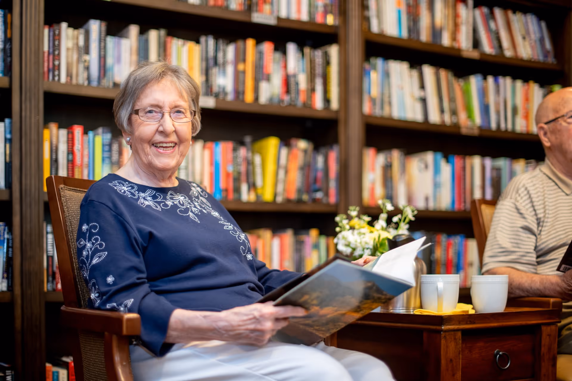 An elderly woman with glasses sitting in a wooden chair in a library or reading room, smiling and holding an open book. Behind her are shelves filled with books. A man is partially visible sitting next to her, and there are two white mugs and a small flower arrangement on a wooden table in front of them.