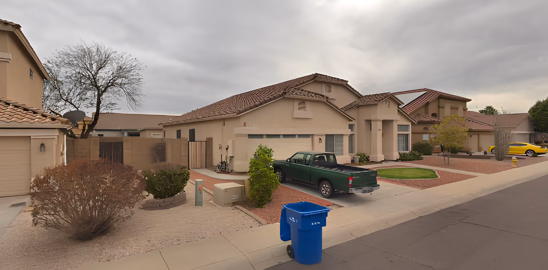 Exterior view of a residential neighborhood street with single-story houses featuring tiled roofs, a green pickup truck parked in a driveway, a blue recycling bin on the curb, and some trees and shrubs in the yards under a cloudy sky.