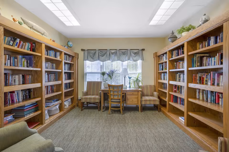 Sunlit library/reading room with wooden bookshelves lining both walls, a desk and chairs by a window.