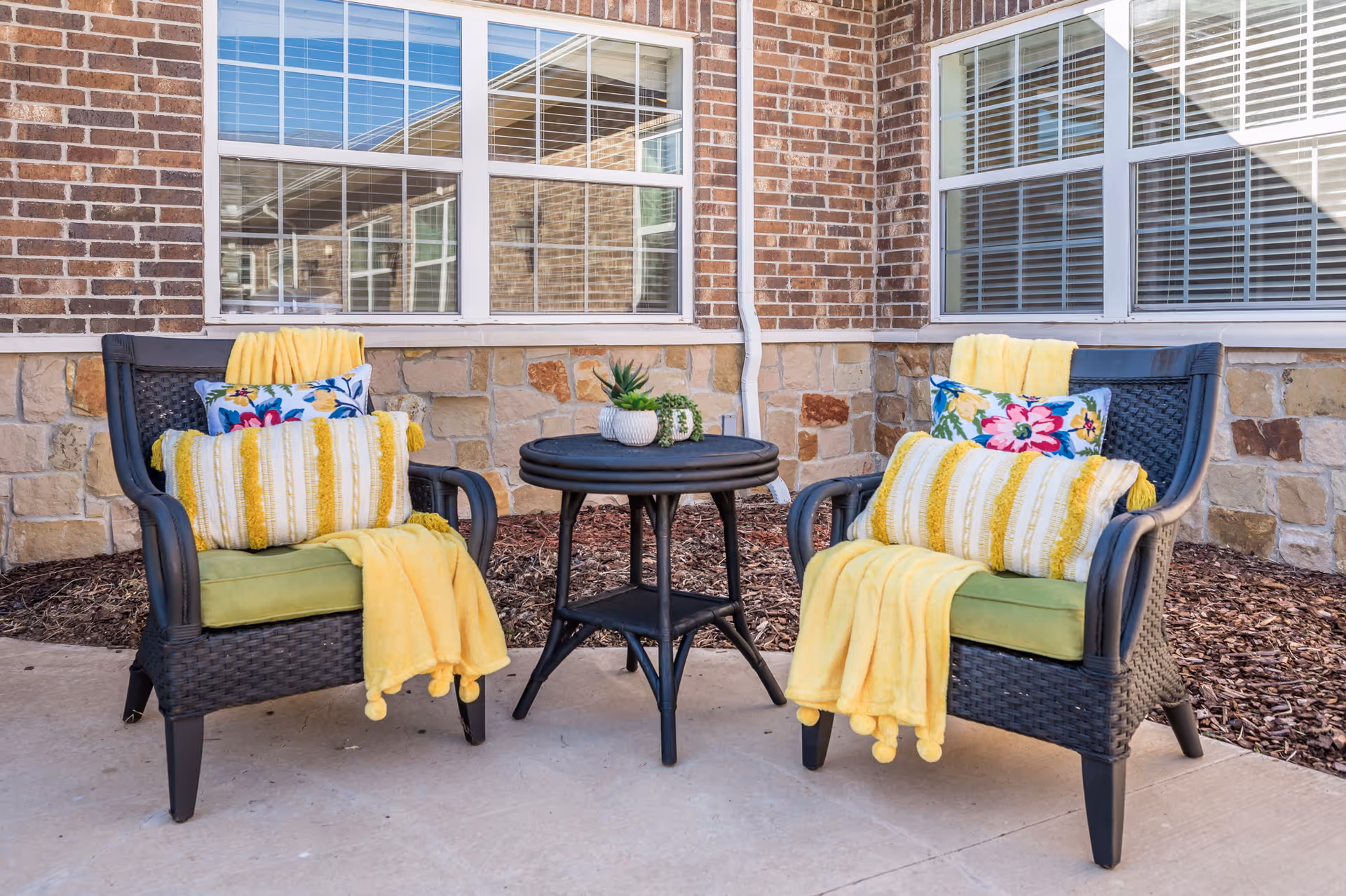 Two black wicker chairs with green cushions, yellow blankets, and decorative pillows with floral and striped patterns are arranged around a small black round table with potted plants on top. The setting is outdoors on a concrete patio in front of a brick and stone building with windows.