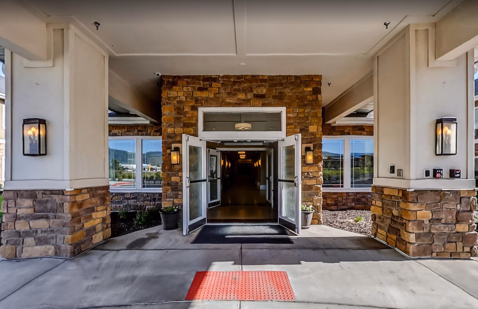 Covered main entrance to a senior living facility with double glass doors set in a stone facade, flanked by columns and wall lanterns.
