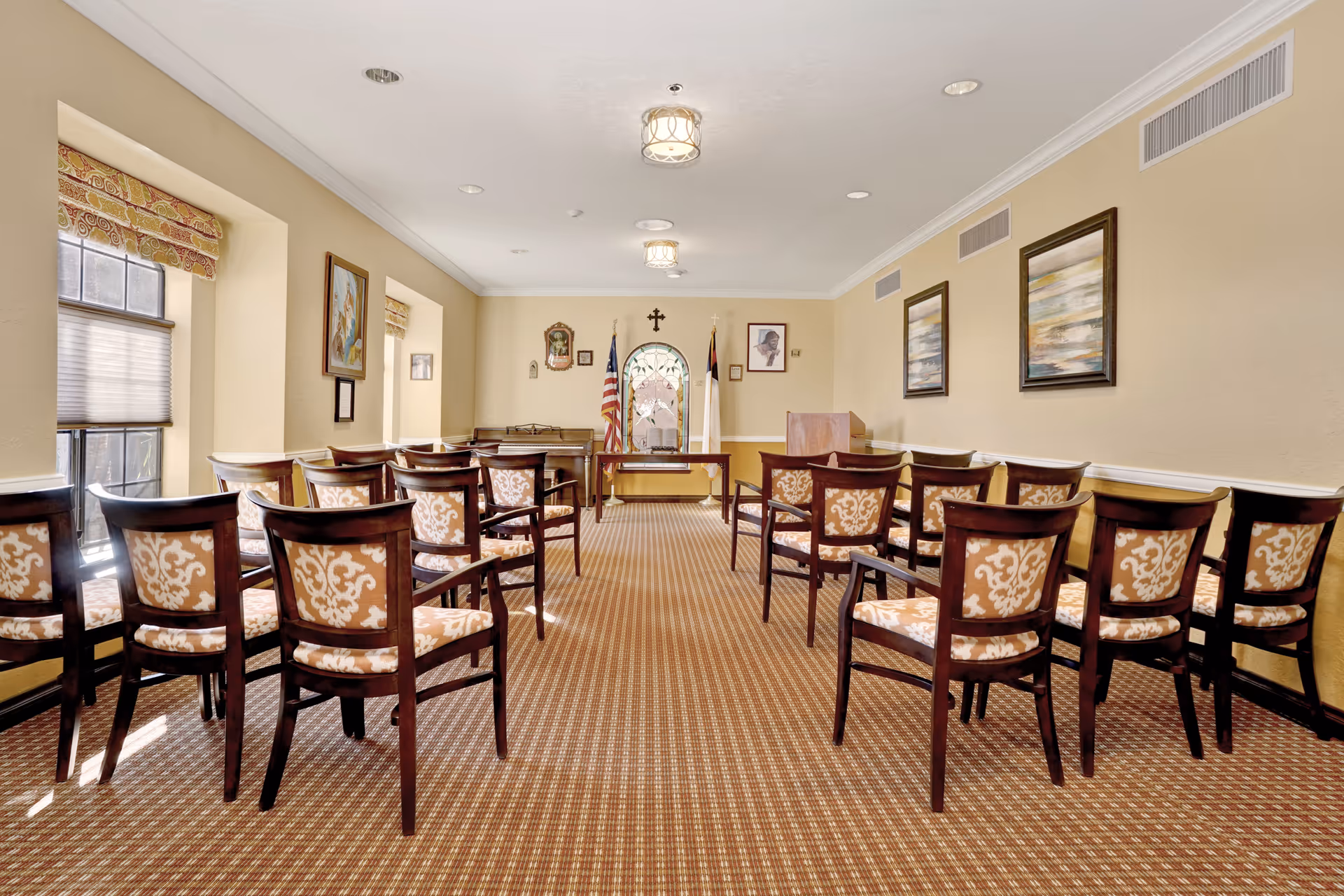A small chapel or meeting room with rows of wooden chairs with patterned upholstery facing a small altar area. The room has beige walls, carpeted floor, framed artwork on the walls, and two flags behind the altar. There is a stained glass window and a wooden podium near the front.