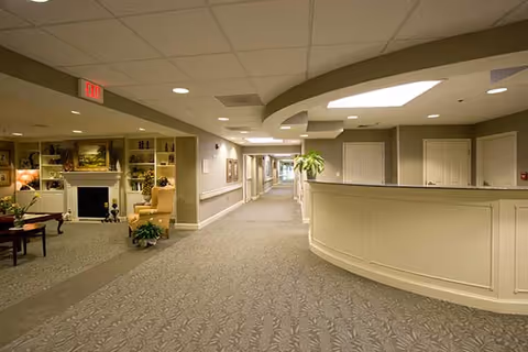 Interior view of a nursing center hallway with a reception desk on the right and a sitting area with chairs and a fireplace on the left. The hallway is carpeted and well-lit with ceiling lights and has handrails along the walls.