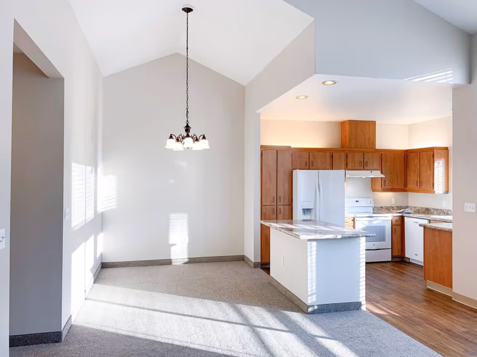 Bright interior space featuring a kitchen with wooden cabinets, white appliances including a refrigerator, stove, and dishwasher, and a marble countertop island. Adjacent to the kitchen is an empty dining area with a hanging light fixture and carpeted floor. Sunlight streams through windows, casting shadows on the walls and floor.