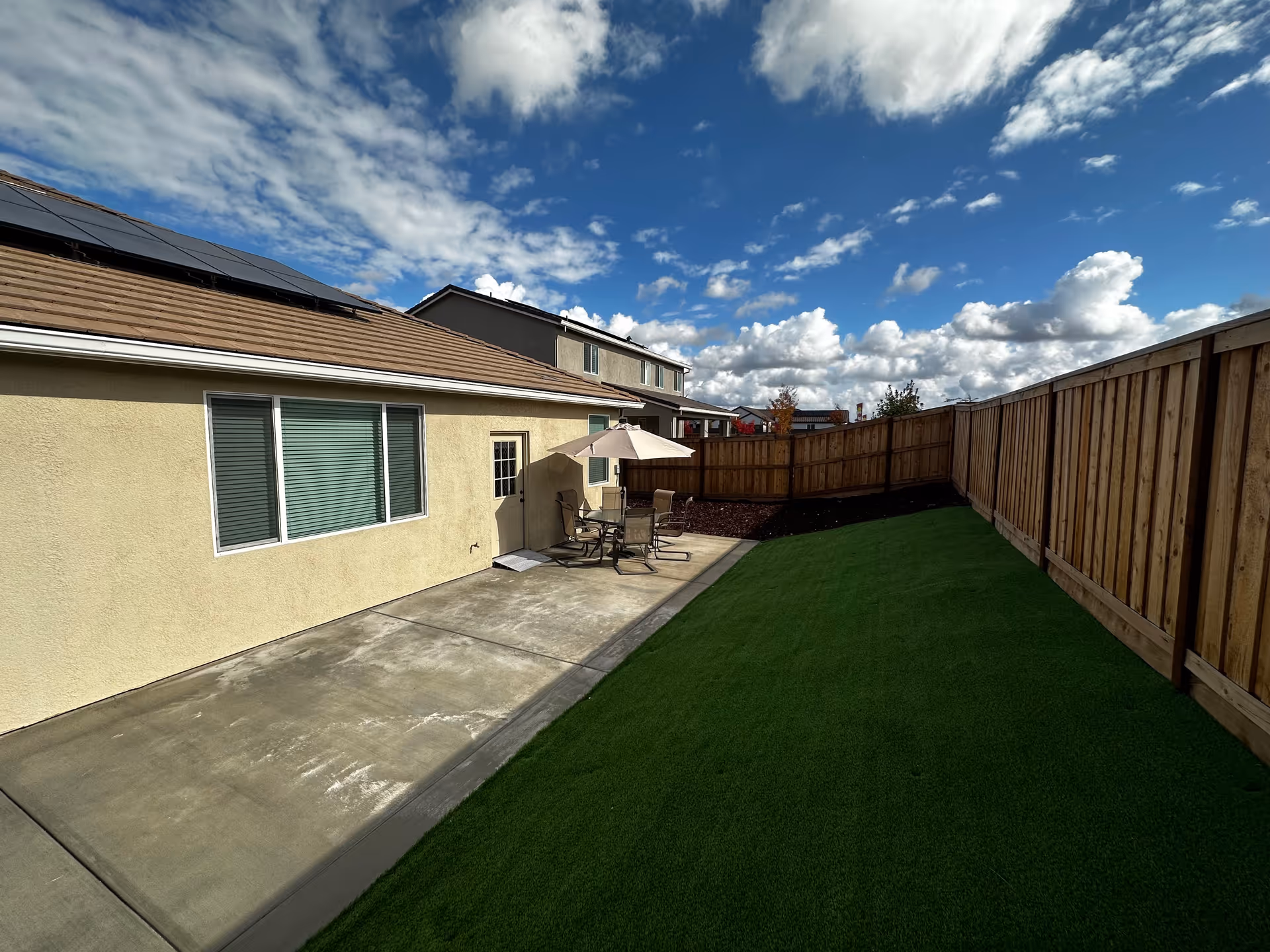 Outdoor patio area with a concrete walkway and a small seating arrangement consisting of a table, four chairs, and a large umbrella. The patio is adjacent to a beige building with windows and a door. The area is enclosed by a wooden fence, and there is a green artificial turf lawn beside the patio. The sky is partly cloudy with patches of blue.