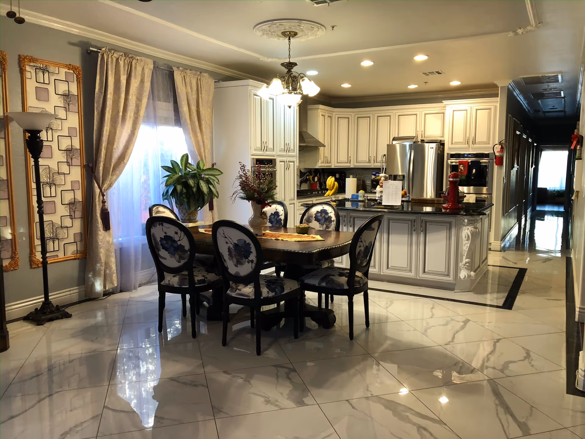 A bright and elegant kitchen and dining area with white cabinetry, a stainless steel refrigerator, and a kitchen island with a black countertop. The dining table has six chairs with floral upholstery. There are large windows with sheer curtains and decorative drapes, a floor lamp, and a plant on the table. The floor is covered with glossy white tiles with gray veining.